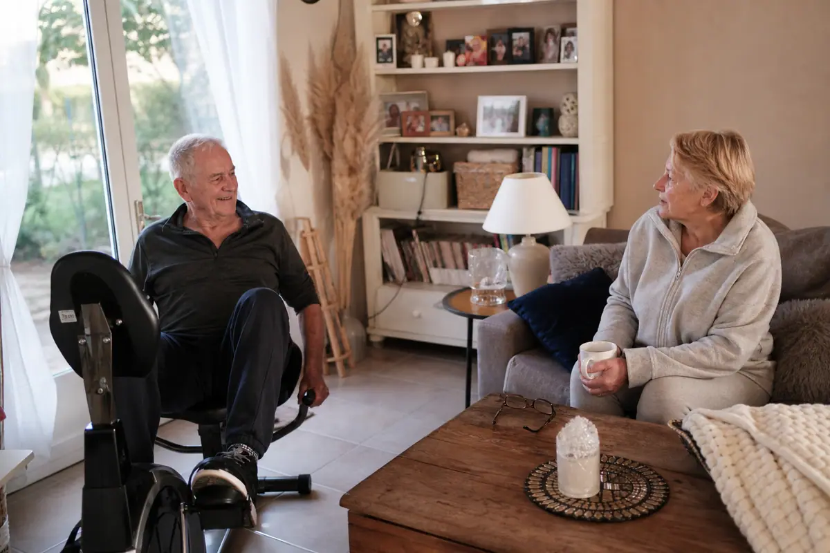 Un retraité pédale sur un vélo semi-allongé dans son salon pendant que son épouse discute avec lui