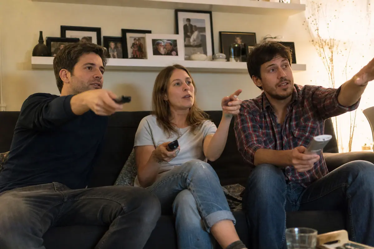 Trois frères et sœurs comparent les télécommandes de différentes box de streaming sur leur canapé