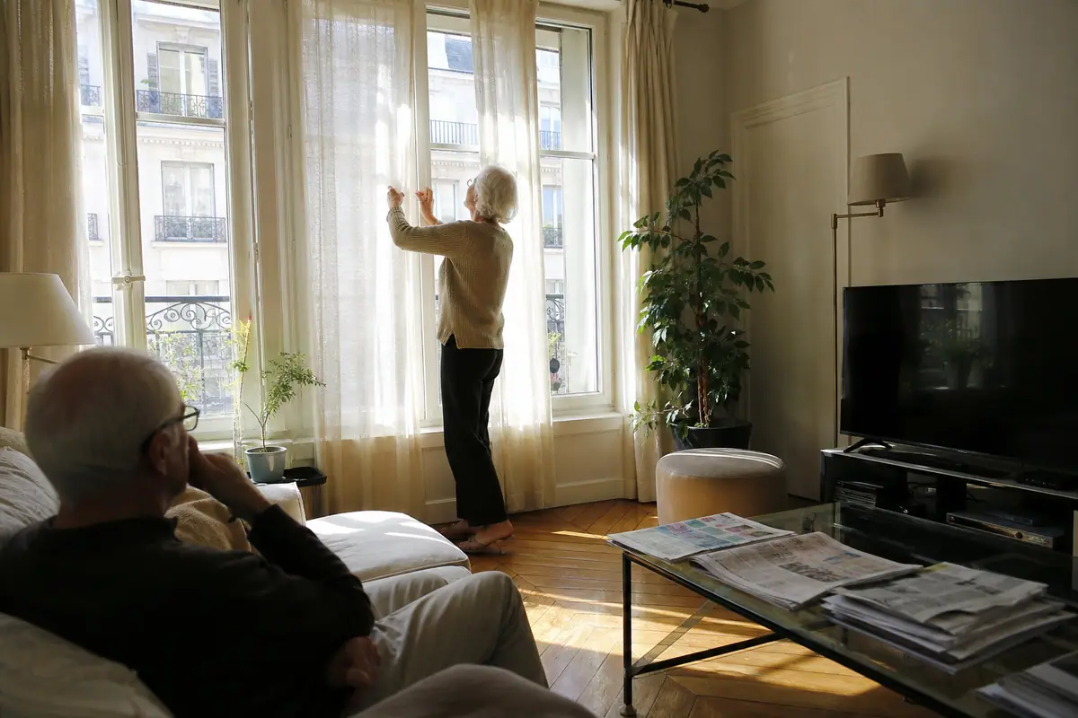 Une femme tire les rideaux d'un salon très lumineux pendant que son mari regarde la télévision
