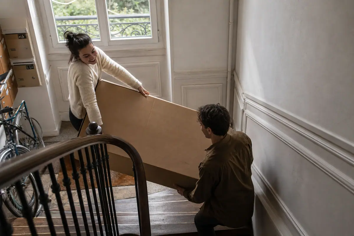 Un jeune couple monte un grand carton de téléviseur dans l'escalier étroit de son immeuble