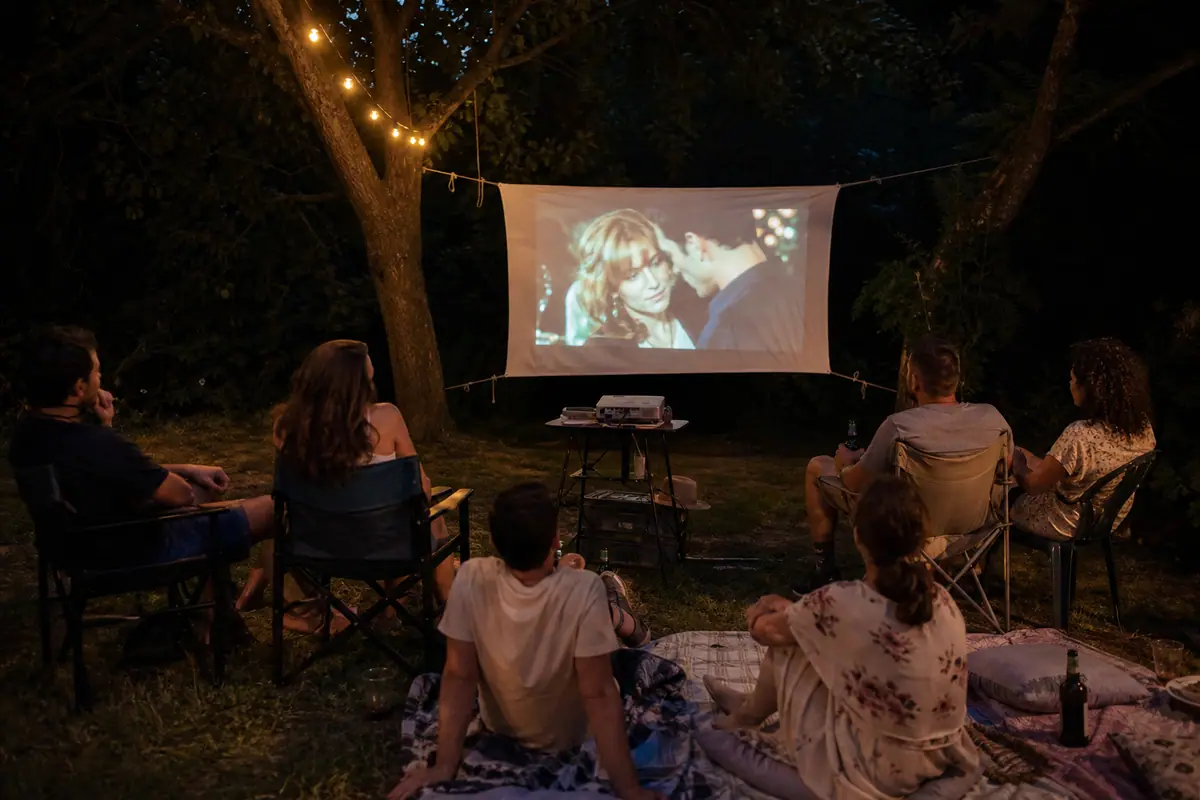 Un groupe d'amis regarde un film projeté sur un drap tendu entre deux arbres lors d'une soirée cinéma en plein air