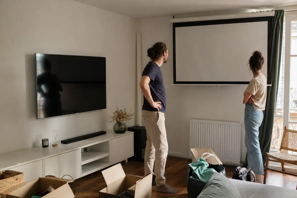 Un jeune couple compare un téléviseur mural et un écran de projection enroulé dans leur salon en cours d'installation